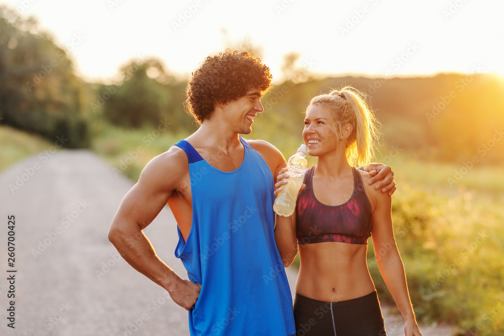 Muscular man standing and hugging his girlfriend while she looking a him and drinking water. Rural exterior, sunny summer day.