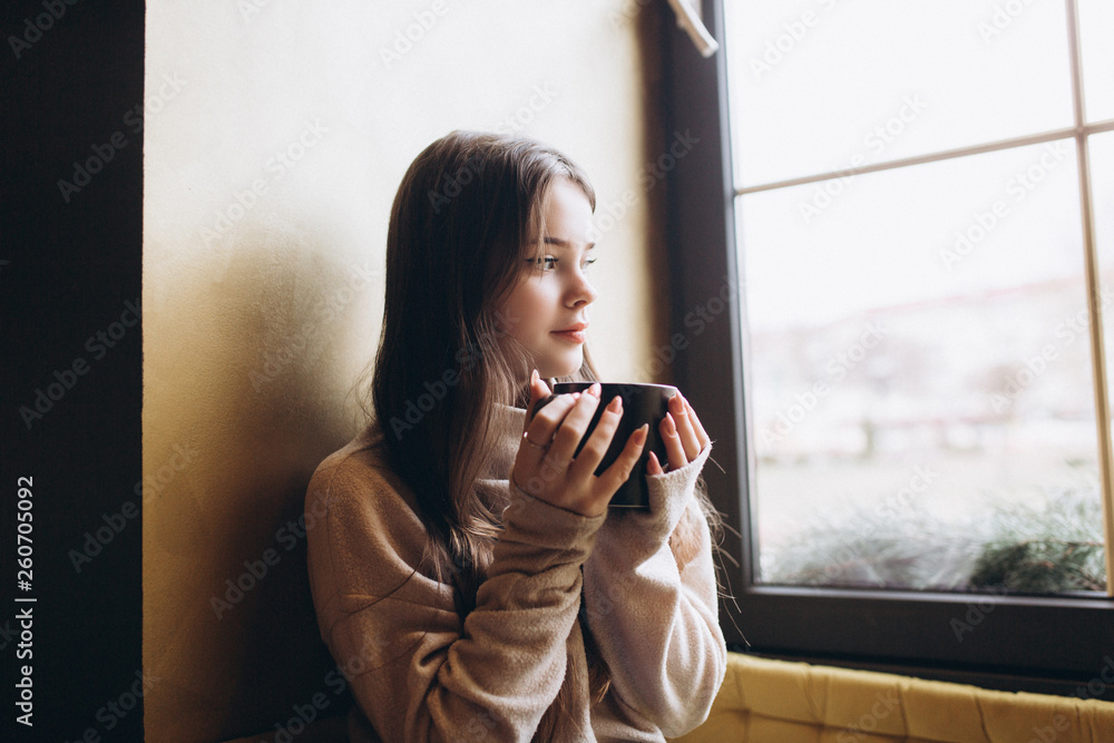 Cheerful young girl having breakfast at cozy home with big cup of coffee or tea holding in hands. Lifestyle, relaxation, natural beauty concept Cheerful young girl having breakfast at cozy home with big cup of coffee or tea holding in hands. Lifestyle, relaxation, natural beauty concept