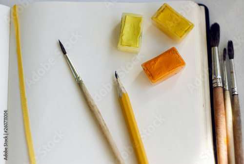On an open notebook with a yellow bookmark are watercolor paints in cuvettes, brushes from squirrel hair and a simple pencil for drawing. Everything is ready for creativity.