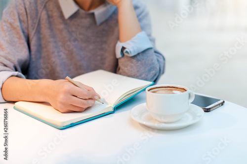 Young woman is writing in notebook in cozy cafe with cup of coffee. Freelancer is working in comfortable coworking modern place. Girl is planning and dreaming. Wellbeing in workplace.