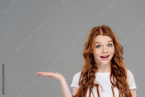 surprised redhead girl gesturing with hand and presenting something isolated on grey with copy space