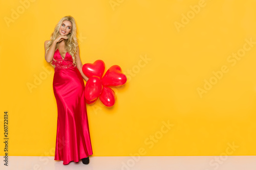 Beautiful Woman In Elegant Red Dress Is Holding Heart Shaped Balloons, Smiling And Looking Away