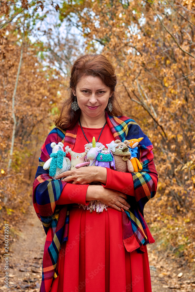 Woman in a red dress with many handmade textile soft toys animals in hands. Bear, owl, unicorn, fox, bunny. The concept of peace, childhood, joy, happiness and pleasure