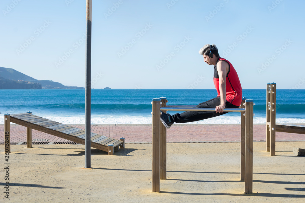 Young boy, practicing sports, calisthenics, with his headphones, outdoors, near the coast, with the blue sea in the background.