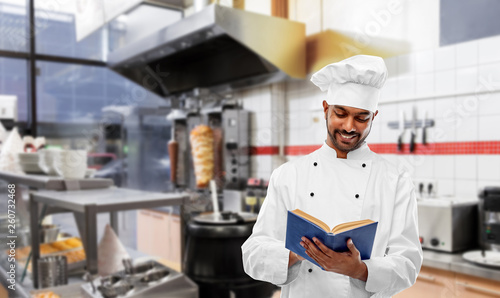 cooking, profession and people concept - happy male indian chef in toque reading cookbook over kebab shop kitchen background