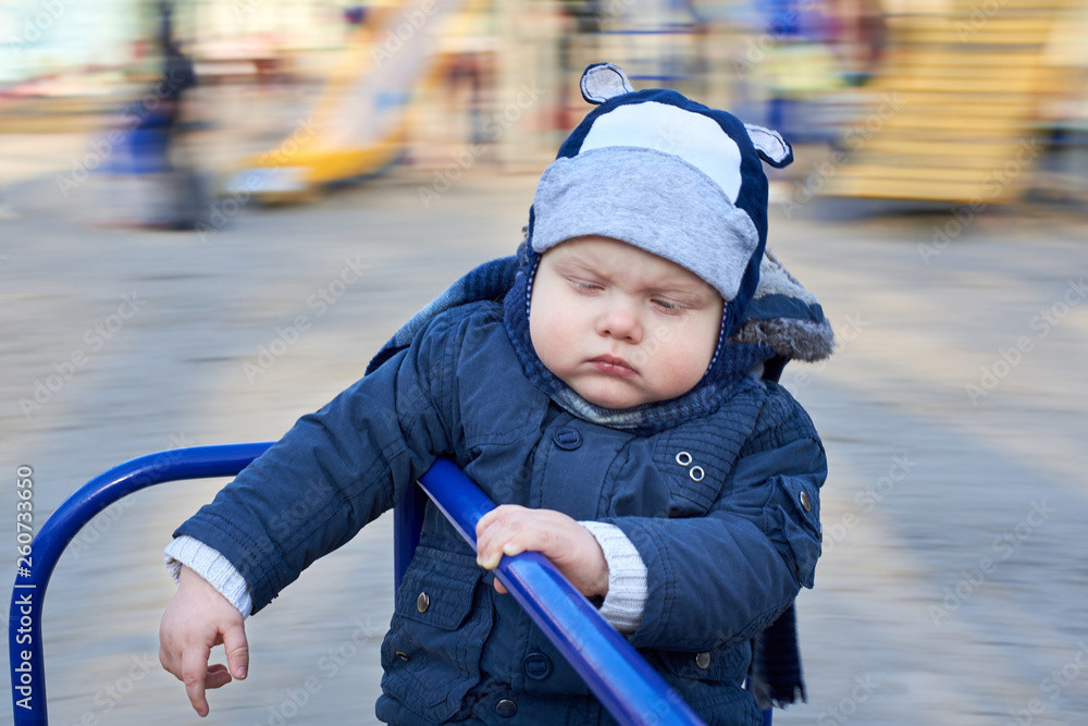 A gloomy pensive little boy rides a merry-go-round on the playground