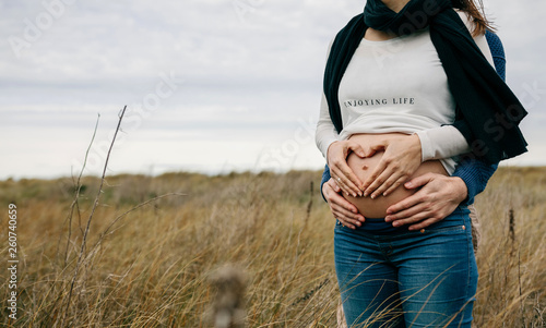 Pregnant making a heart with hands on her naked belly while her partner embraces her photo