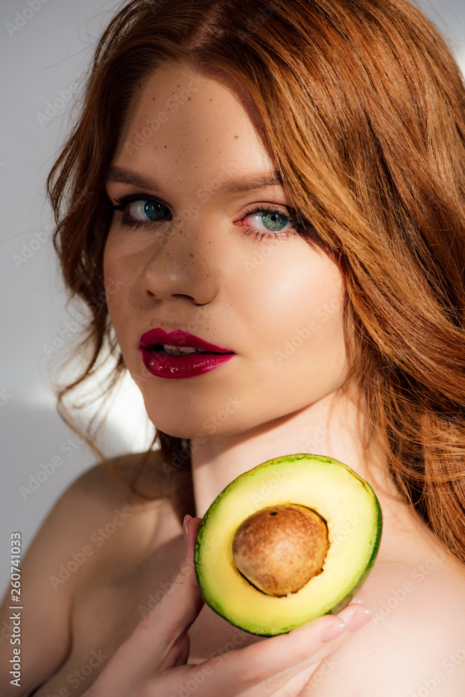 beautiful redhead girl with red lips looking at camera and posing with cut avocado beautiful redhead girl with red lips looking at camera and posing with cut avocado