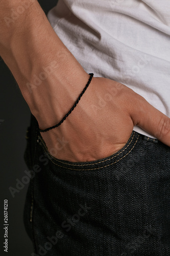 Cropped closeup shot of man's hand with tanned skin, wearing black lucky rope bracelet. The guy is wearing black jeans and white shirt, putting his hand into the pocket, posing on dark background.