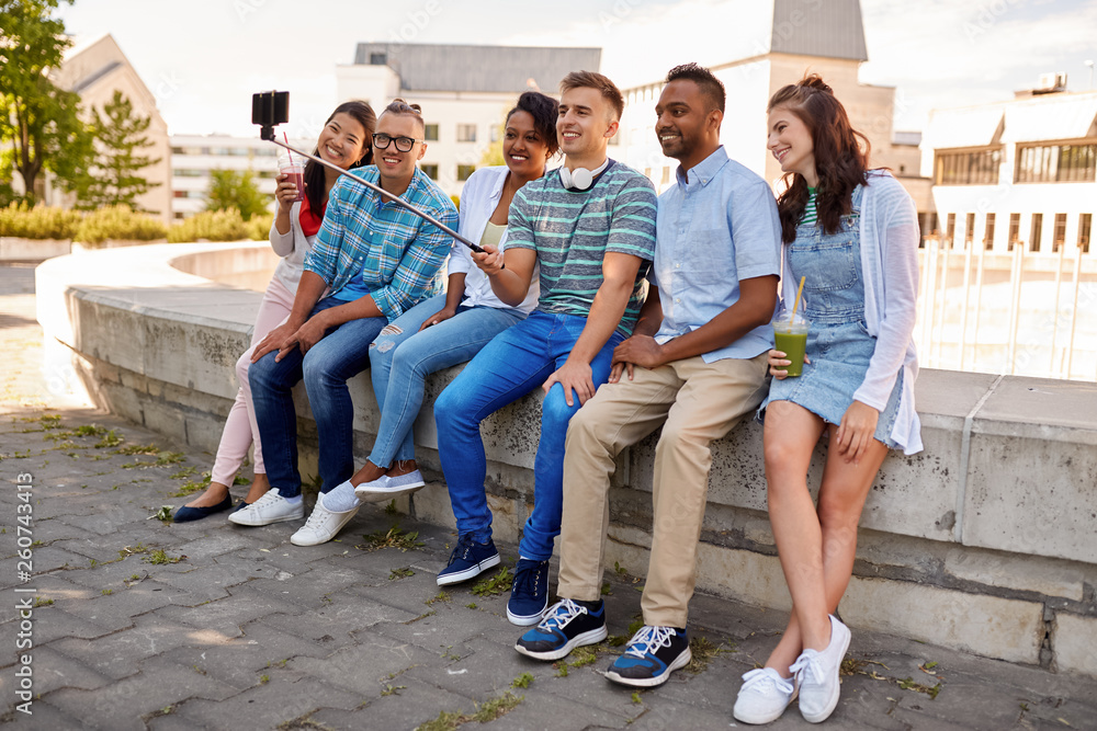 Fototapeta premium people, technology and friendship concept - group of happy international friends or students taking picture by smartpnone on selfie stick in city