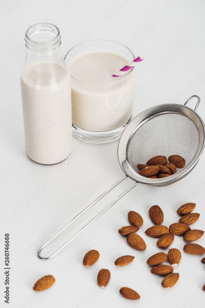 vegan almond milk in bottle and glass with straw near scattered almonds and sieve vegan almond milk in bottle and glass with straw near scattered almonds and sieve