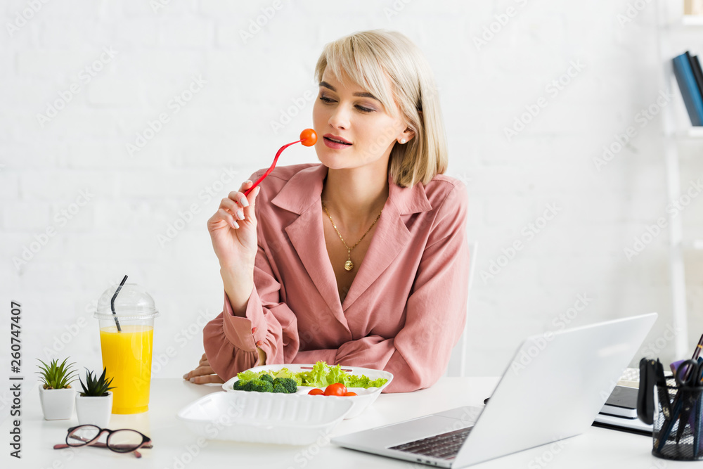 blonde freelancer holding fork with cherry tomato near laptop blonde freelancer holding fork with cherry tomato near laptop