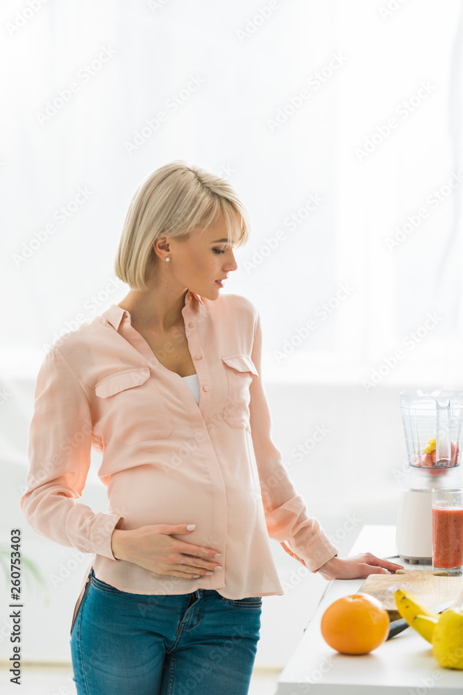 blonde pregnant woman looking at glass of smoothie in kitchen