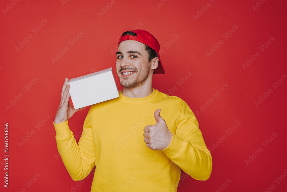 Young guy in cap holding a box and showing thumb up Young guy in cap holding a box and showing thumb up