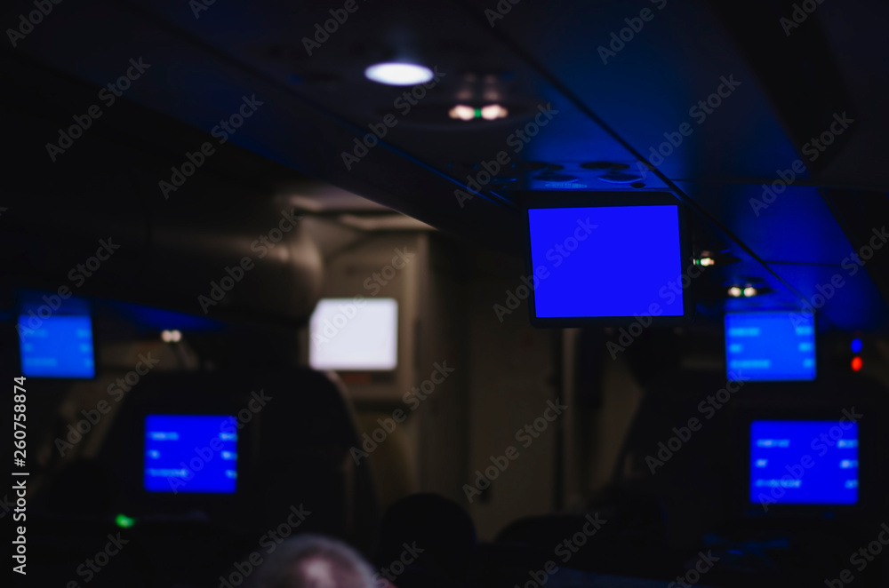 Inside an airplane during a flight with the informative tv screens down. Plane with low light, tvs with a informative blue screen.