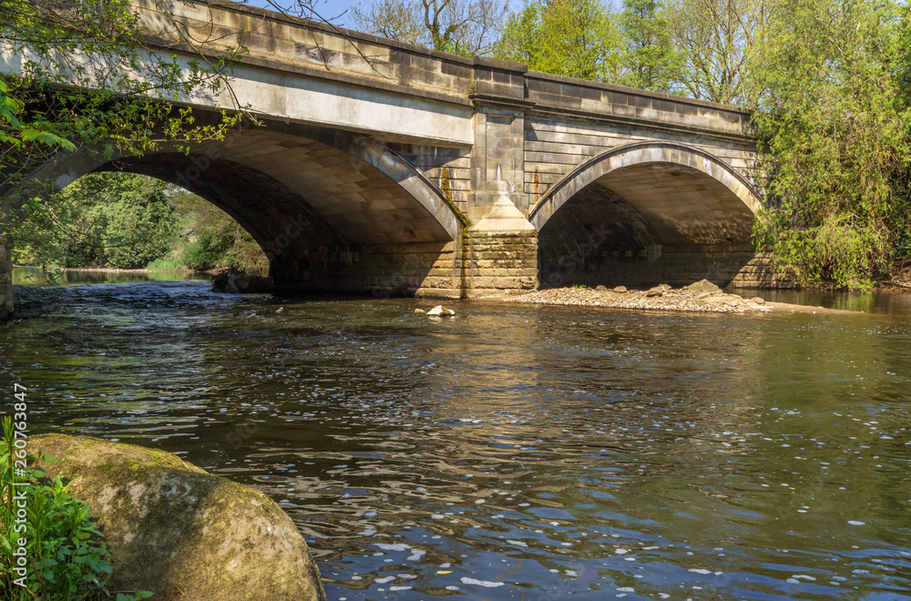 A stone bridge arches over a tranquil flowing river on an idyllic summers day