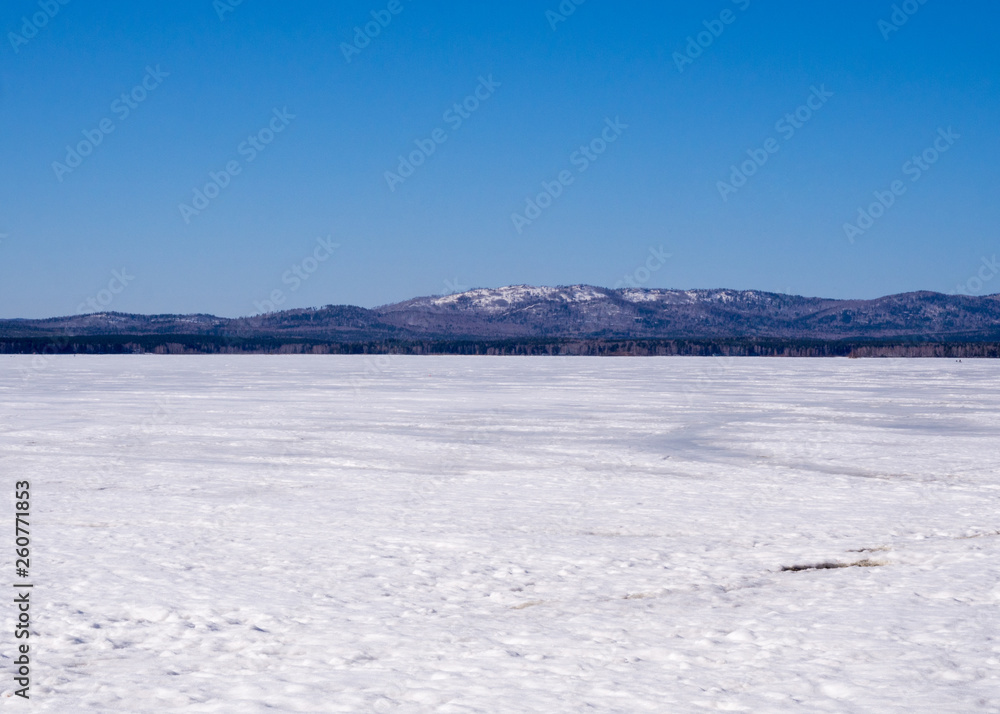 Winter landscape, frozen lake under snow and mountains on a skyline. Blue sky, sunny cloudless day.