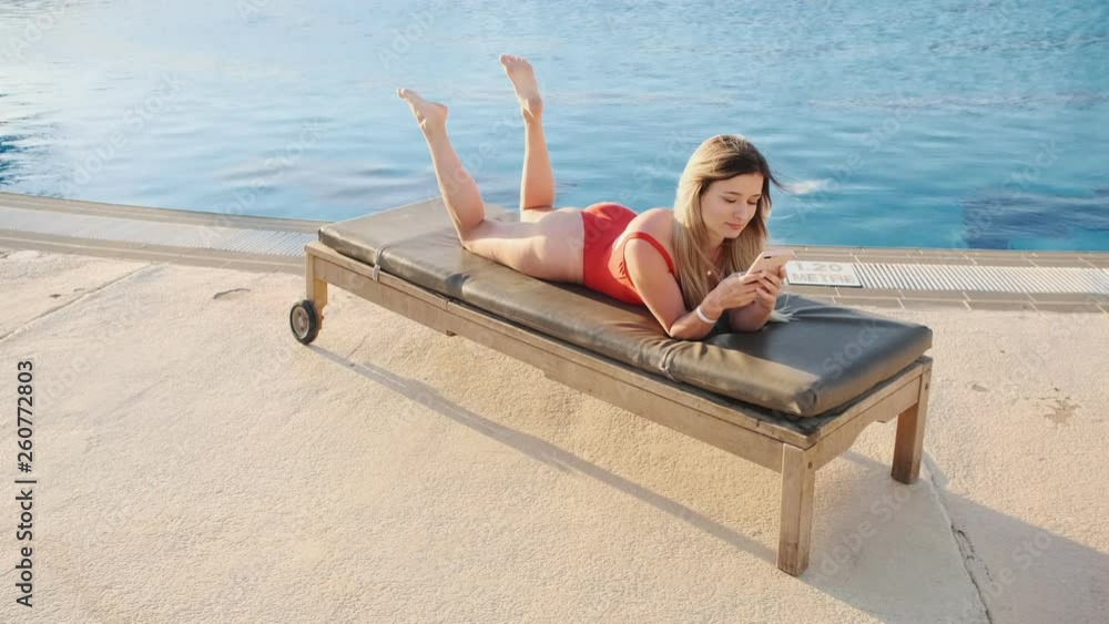 Top view of stylish sexy woman in red bikini lying on deck chair, using smartphone app, browsing the internet, texting in hotel pool side area. Girl blogging online and sunbathing, relaxing at resort.