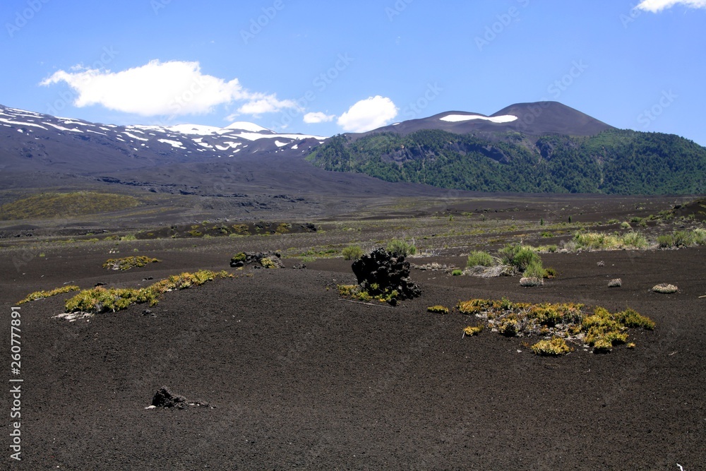 View over wide field of volcanic lava ash on peak of black Volcano Llaima with spots and stripes of snow and ice contrasting with blue sky - Conguillio NP in central Chile