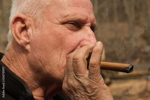 Portrait of a man with a weathered faace puffing a cigar with his working hands showing photo