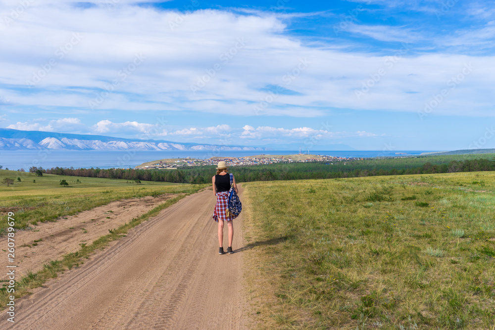 Tween tourist girl in hat and backpack walking on country road alone and admiring picturesque landscape of lake Baikal, mountains, village and blue sky. Travelling, hiking and summer vacation concept