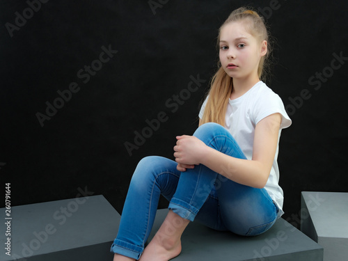 A concept portrait of a cute pretty blonde teen girl with long hair sitting in a white t-shirt against a black background in the studio and talking.