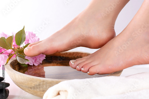 Beautiful female feet in spa studio with french nails polish. Girl legs on wooden bowl with water and pink flowers and towel isolated on white background. Close up, selective focus