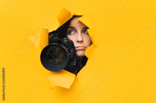 A young paparazzi girl holds a reflex camera and looks through a torn hole in yellow paper. The concept of embarrassment, sadness, disappointment, despondency and upset. Copy space.