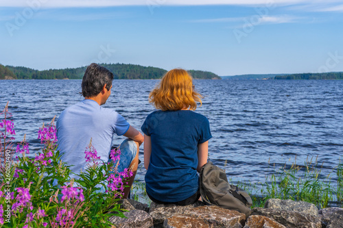 Tourists dark-haired man and redhead young woman sitting on stones on shore of lake bay in sunny summer evening. People resting and admiring beautiful landscape of Ladoga lake. Karelia, Russia