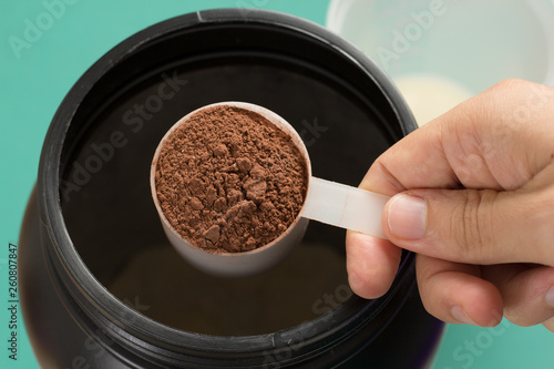 Whey Protein. Point of view of hand holding measuring scoop. Chocolate flavour. Color background: green.