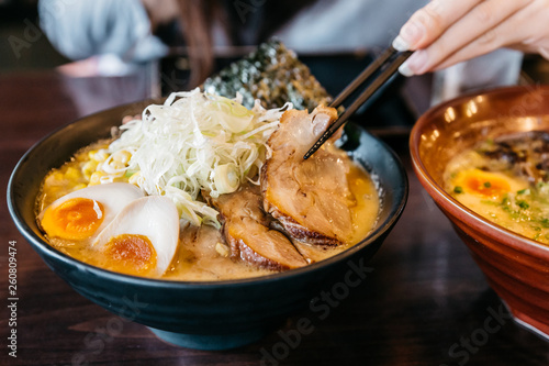 Woman hand pinching noodle in Ramen Pork Bone Soup (Tonkotsu Ramen) with Chashu Pork, Scallion, Sprout, Corn, Dried Seaweed and boiled egg served in black bowl.