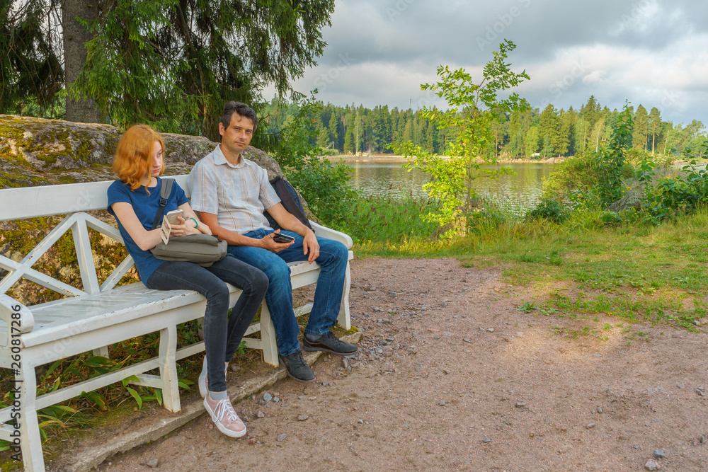 Handsome middle-aged man and young pretty lady sitting on bench and using smartphones in summer evening. Tourists on the beautiful landscape background. Monrepos Park, Vyborg, Russia. Travel concept
