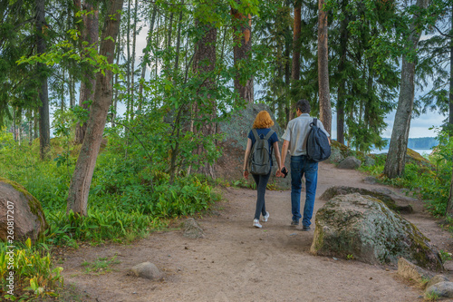 Dark-haired middle-aged man and red-haired young lady walk along forest road. Tourists on the beautiful landscape background. Monrepos Park, Vyborg, Russia. Travel concept