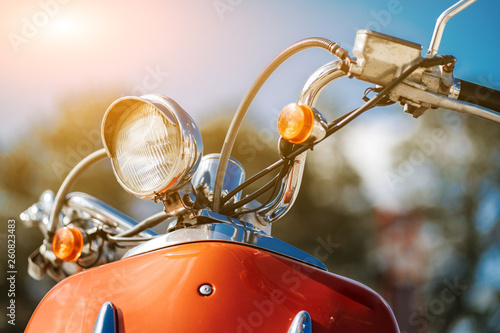 Closeup chrome detail and headlamp of orange retro vintage scooter under blue sky and sun on the blurred background.