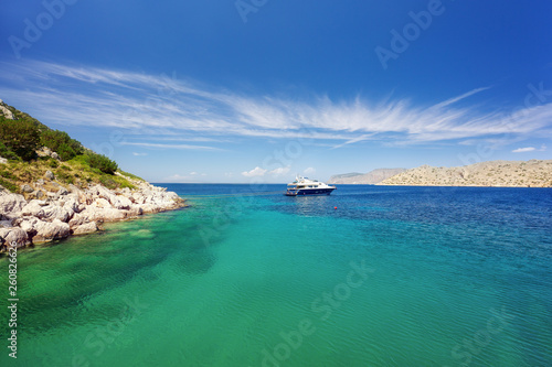 Crystal clear turquoise water in a lagoon near Bisti Beach on Hydra Island Greece. Unrecognizable people relaxing on a luxury yacht with mooring ropes against the rocks