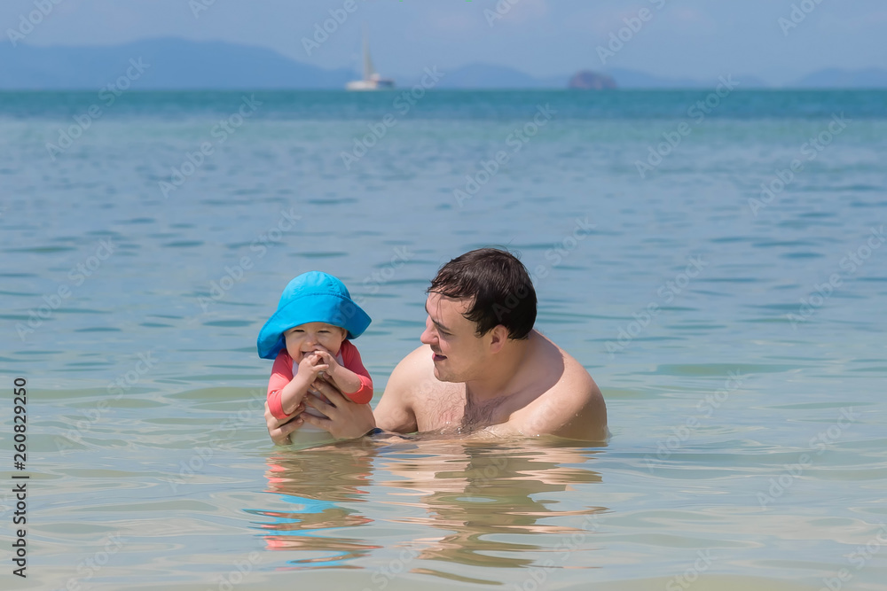 Infant baby girl first time in the sea. She is very impressed and happy but teething. Father holds smiled child in the water and teaches to swim. Summer vacation on tropical beach