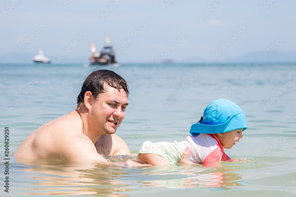 Father holds smiled and happy child in the water and teaches to swim. Summer vacation on tropical beach. Infant baby girl 9 month first time in the sea
