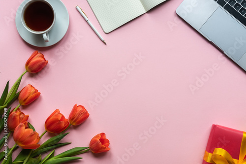 Red tulips with cup of tea, laptop, gift box and notebook on pink background. Copy space