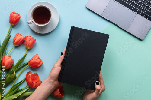 Hands holding book on blue background. Background with coffee, laptop and flowers