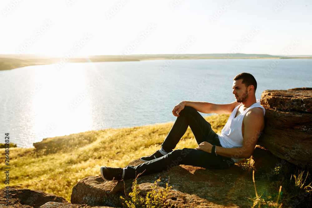 Handsome young man sitting on lawn nexto to a lake in a sunny, peaceful day, looking away to a side