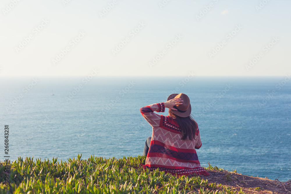 Closeup back view of woman in travel clothes and hat sitting and looking at blue ocean and sky.