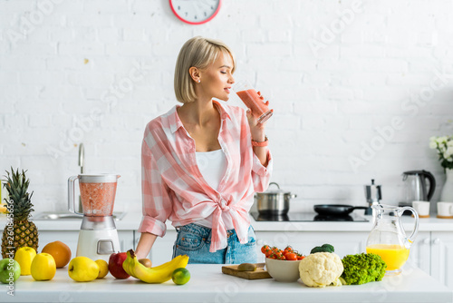 Fotografie attractive blonde woman drinking tasty smoothie near ingredients in kitchen