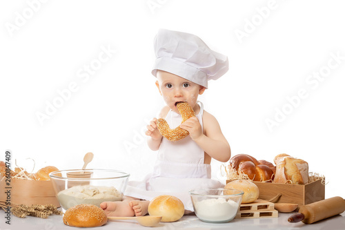 The boy in the costume of the chef in front of the basket with bread and with the bagel in hand. Cooking child lifestyle concept. Toddler playing