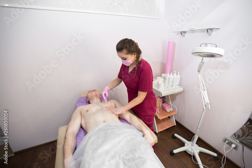Master depilation works with a client man in a beauty salon  it removes hair from the mans chest. A young guy is lying on the table in the office of hair removal