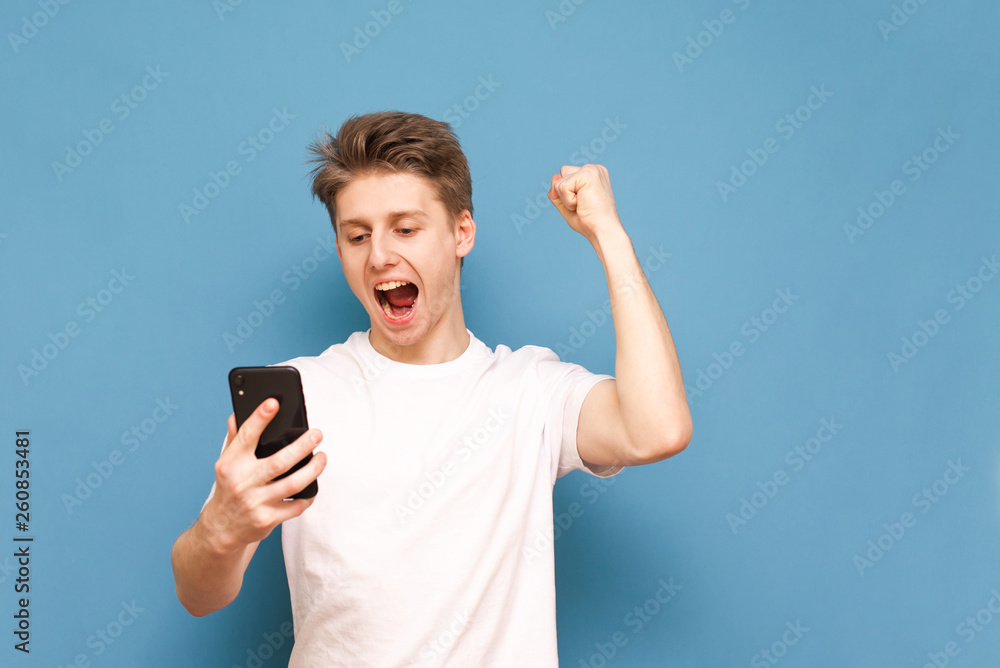 Portrait of a joyful guy in a white T-shirt, rejoices in victory and looks at the screen of a smartphone on a blue background. Joyful guy is using a smartphone and is glad, isolated. Portrait of a joyful guy in a white T-shirt, rejoices in victory and looks at the screen of a smartphone on a blue background. Joyful guy is using a smartphone and is glad, isolated.