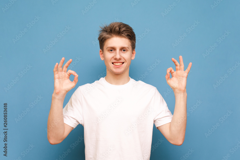 Smiling guy in a white T-shirt shows an OK gesture with his hands and looks at the camera on a blue background. Happy young man shows an OK sign, isolated. Smiling guy in a white T-shirt shows an OK gesture with his hands and looks at the camera on a blue background. Happy young man shows an OK sign, isolated.