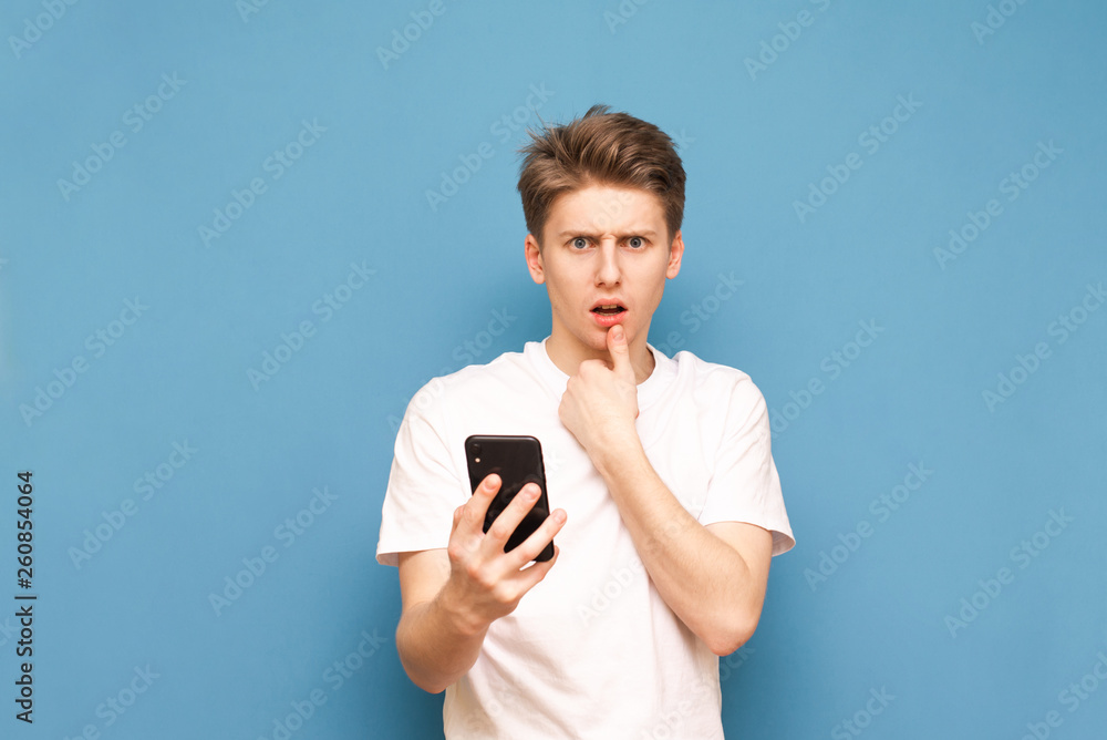 Portrait of a surprised young man standing with a smartphone in his hands on a blue background, looking into the camera. Puzzled guy in a white T-shirt with a smartphone in his hand, isolated Portrait of a surprised young man standing with a smartphone in his hands on a blue background, looking into the camera. Puzzled guy in a white T-shirt with a smartphone in his hand, isolated