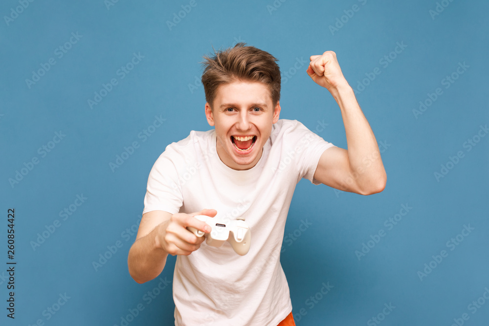Naklejka premium Portrait of a happy gamer plays video games, rejoices in victory on a blue background, looks into the camera and smiles. Joyful gamer with a joystick in his hand is isolated on a blue background.