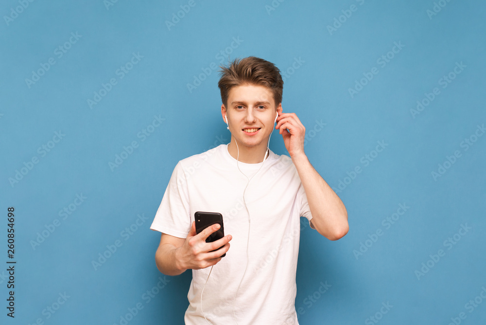 Cheerful guy standing with a smartphone in his hand against the background of a blue wall and listening to music in the headphones, wearing a white T-shirt, looking into the camera and smiling. Cheerful guy standing with a smartphone in his hand against the background of a blue wall and listening to music in the headphones, wearing a white T-shirt, looking into the camera and smiling.