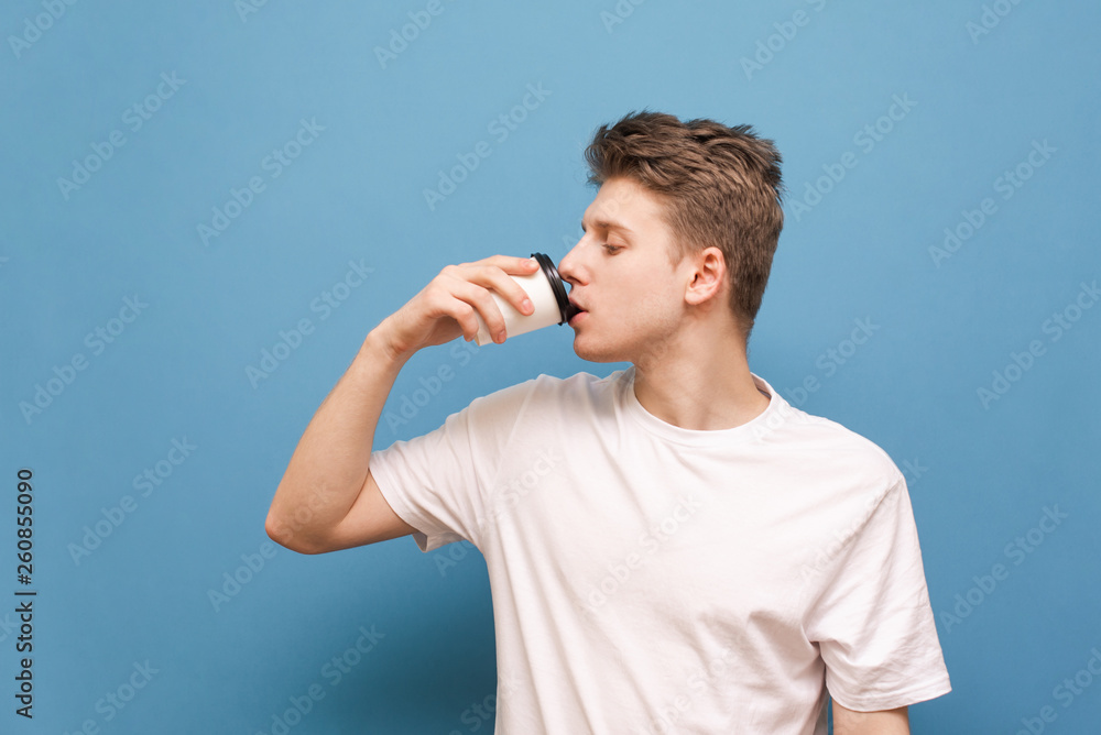 Young man in a white T-shirt drinking coffee from a paper cup on a blue background looks to the side. Isolated The guy drinks a hot drink from a paper cup. Copyspace Young man in a white T-shirt drinking coffee from a paper cup on a blue background looks to the side. Isolated The guy drinks a hot drink from a paper cup. Copyspace